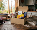 Swedish Baking Box – Small open on a rustic wooden kitchen table showing Dan Sukker pearl sugar, Fazer Ögon Cacao, Dr. Oetker vanilla sugar, KronJäst dry yeast and Santa Maria cardamom, with baking tools and a garden window in the background
