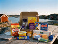 Swedish Breakfast Box Large open on a weathered wooden table by the Swedish west coast archipelago – Ekströms Nyponsoppa, Löfbergs Mellanrost, Havrefras, Kavli Mildost, O'boy Portion, Wasa Frukost, Pågen Hönökaka, Fjällbrynt Rökt Ren, Kalles Kaviar, Fjällbrynt Messmör, Den Gamle Fabrik orange marmalade and Pågen LingonGrova inside, with red boathouses and sea in the background
