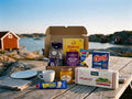 Swedish Breakfast Box Medium open on a weathered wooden table by the Swedish west coast archipelago – Pågen LingonGrova, Havrefras, Löfbergs Mellanrost, Kavli Mildost, Kalles Kaviar, Den Gamle Fabrik orange marmalade, O'boy Portion and Wasa Frukost inside, with red boathouses and sea in the background
