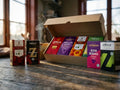 Swedish Coffee Taste Box Medium open on a rustic wooden table in warm morning light, showing 8 Swedish ground coffee bags – Gevalia Mellanrost, Löfbergs Mellanrost Originalet, Arvid Nordquist Gran Dia, Zoégas Fikastund, Zoégas Mollbergs Blandning, Löfbergs Kharisma, Zoégas Skånerost and Arvid Nordquist Franskrost – arranged in and around an open kraft box with a steaming mug in the background
