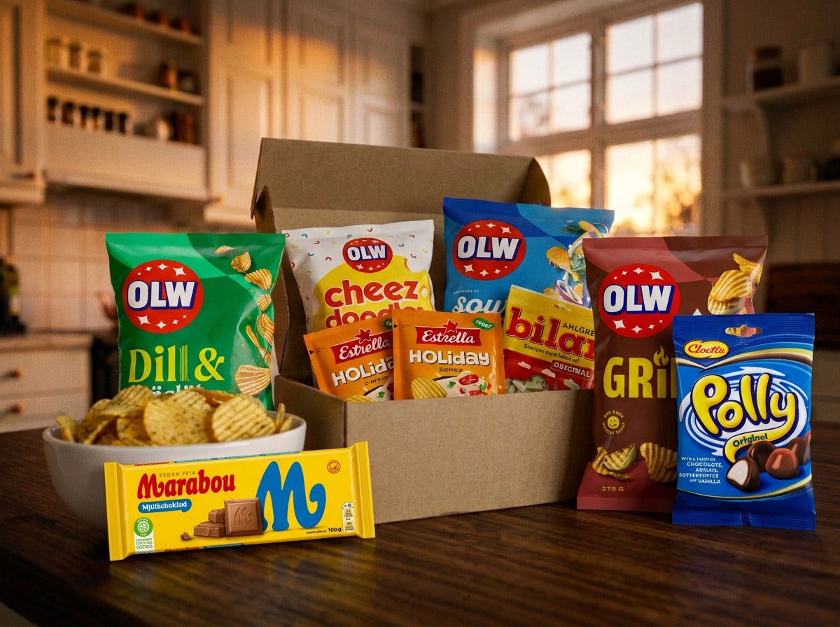 Swedish Fredagsmys Box Medium open on a dark wooden kitchen table in a warm cozy kitchen – OLW Dill & Gräslök, OLW Cheez Doodles, OLW Sourcream & Onion, two Estrella Holiday Dippmix sachets, Ahlgrens Bilar, OLW Grillchips, Cloetta Polly Original and Marabou Mjölkchoklad inside, with a bowl of chips alongside
