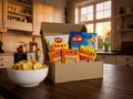 Swedish Fredagsmys Box Small open on a dark wooden kitchen table in a warm cozy kitchen – OLW Cheez Doodles, OLW Sourcream & Onion Chips, two Estrella Holiday Dippmix sachets and Ahlgrens Bilar inside, with a bowl of chips alongside
