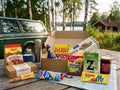Taste of Sweden Box Large open on a wooden table by a Swedish lake next to a vintage Volvo estate – Ekströms Nyponsoppa, Slotts Skånsk Senap, Kavli Mildost, Kalles Kaviar, Ahlgrens Bilar, Wasa Husman, Malaco Djungelvrål, Zoégas Fikastund, lingonberry jam and Marabou Mjölkchoklad inside
