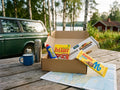 Taste of Sweden Box Small open on a wooden jetty table by a Swedish lake – Kalles Kaviar, Ahlgrens Bilar, Wasa Husman and Marabou Mjölkchoklad inside, with a vintage Volvo and red cottage in the background
