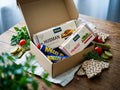 Kalles Kaviar Taste Box Small open on a rustic wooden table – Kalles Original, Kalles Randiga, Wasa Husman and Wasa Frukost crispbread inside, surrounded by cherry tomatoes, cucumber, dill and pickles
